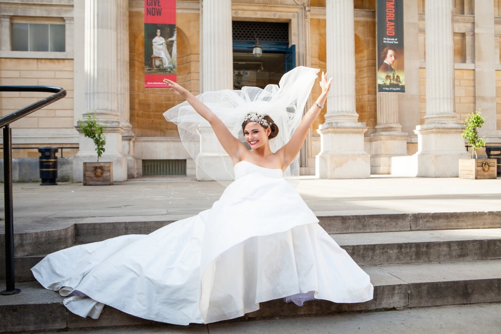 A joyful bride in a white dress and floral headpiece sits on steps outside a grand building, raising her arms and smiling widely. Captured by an Oxfordshire Wedding Photographer, her veil flows behind her as banners hang on the columns. David Bostock Photography