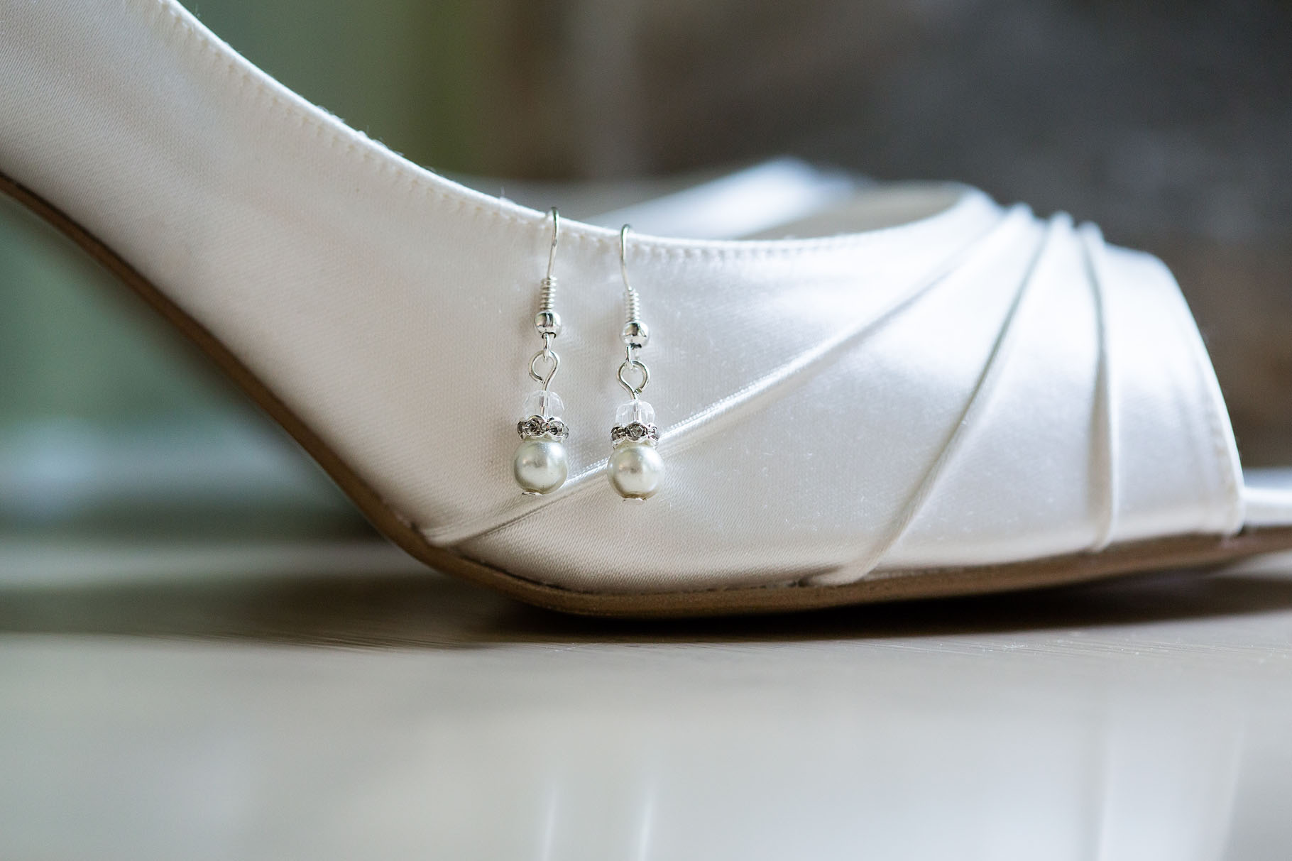 A close-up of a white satin high-heeled shoe with a pair of pearl drop earrings hanging from the heel, elegantly captured by an Oxfordshire wedding photographer on a reflective surface. David Bostock Photography