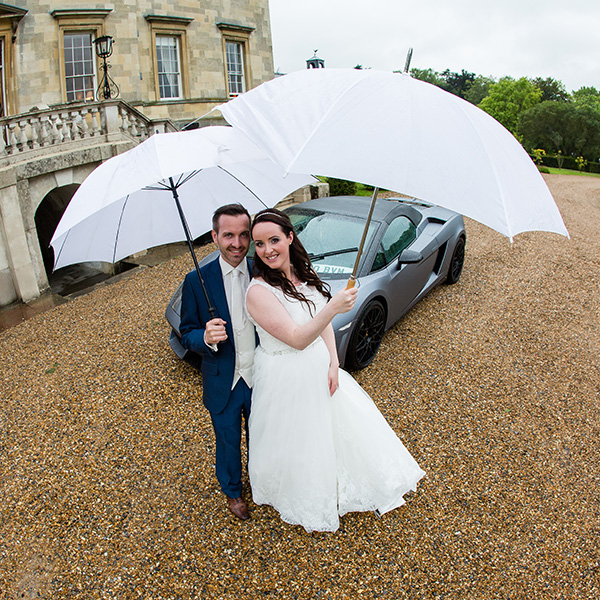 A bride and groom in wedding attire stand on gravel before a large building and a silver sports car, smiling and holding white umbrellas together amidst greenery—capturing the joy seen in David Bostock Photography testimonials. David Bostock Photography