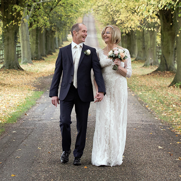 A bride and groom walk hand in hand down a tree-lined path, smiling at each other. The ground is covered in autumn leaves—a scene often praised in David Bostock Photography testimonials for its beauty and emotion. David Bostock Photography