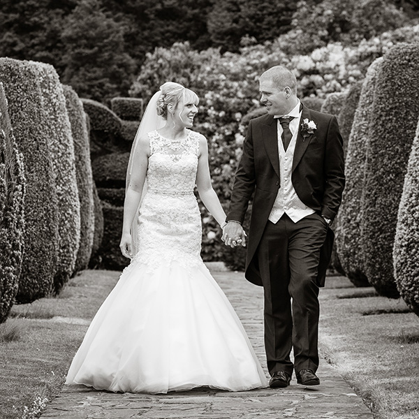 A bride and groom hold hands and smile at each other whilst walking along a stone path in a manicured garden, surrounded by sculpted bushes. This black and white moment is often praised in David Bostock Photography testimonials. David Bostock Photography