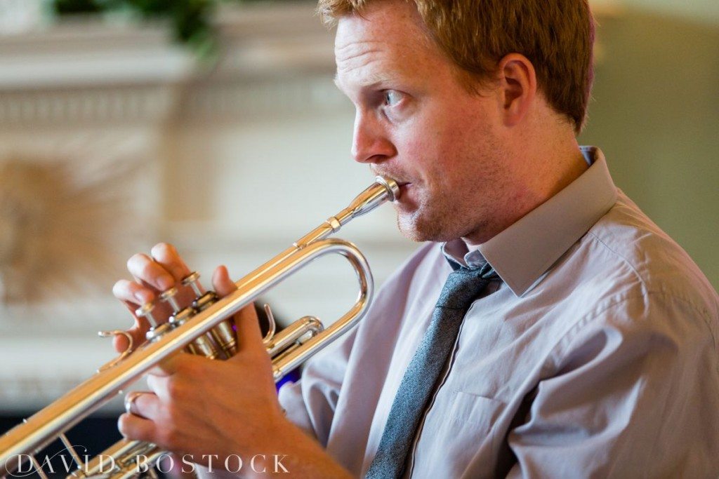 A man wearing a light purple shirt and grey tie plays a silver trumpet indoors at a Kirtlington Park wedding, focusing intently on his performance. David Bostock Photography