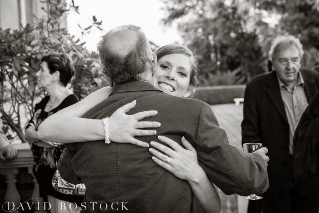 A smiling woman hugs a man, her face beaming and partially visible over his shoulder. She holds a wine glass, with several guests mingling in the background at a joyful Kirtlington Park wedding. David Bostock Photography