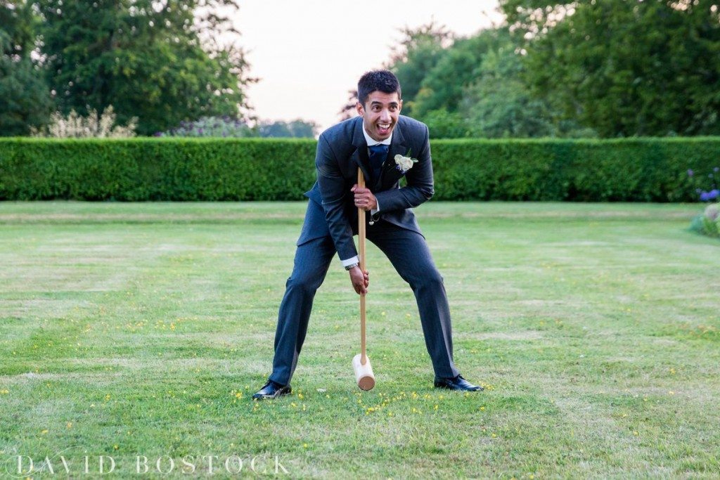A man in a suit, smiling and holding a croquet mallet, stands on a grassy lawn surrounded by trees and bushes at a Kirtlington Park wedding. David Bostock Photography