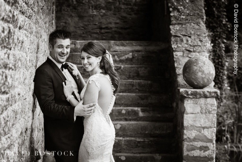 A bride and groom stand smiling and embracing next to a stone wall and staircase at Le Manoir. The bride wears a lace dress, and the groom is in a tuxedo. The rustic setting features textured stone and a round ornament on the ledge. David Bostock Photography