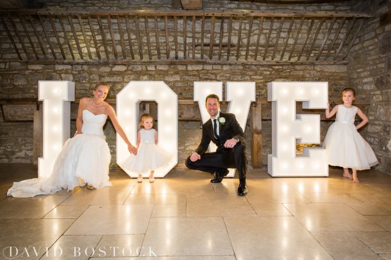 A bride, groom, and two young girls in white dresses pose and smile in front of large, illuminated LOVE letters against a rustic stone wall, captured beautifully by a Caswell House Wedding Photographer. David Bostock Photography
