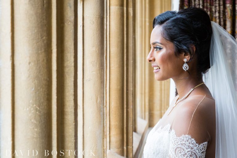 A bride in a white lace off-shoulder gown and veil smiles whilst looking out of tall stone columns, wearing pearl jewellery. Sunlight illuminates her face. David Bostock Photography