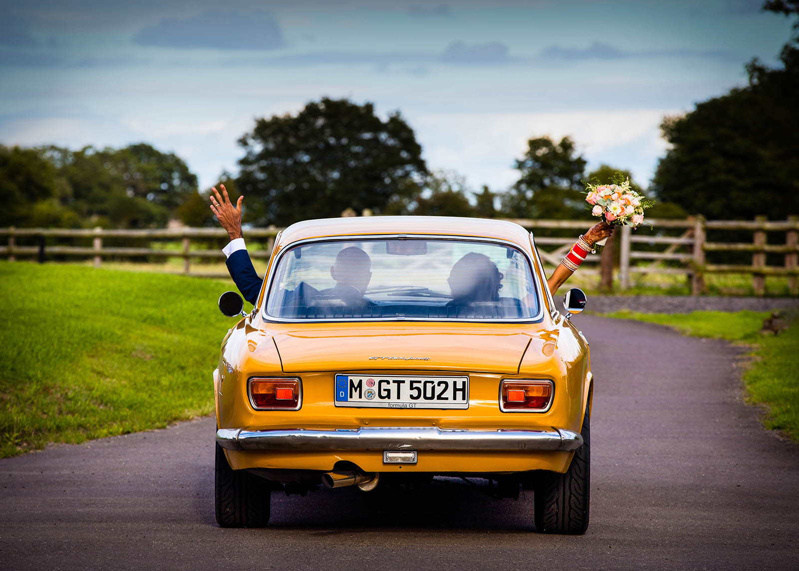 Bride and Groom driving away in a orange car with their arms out the window waving