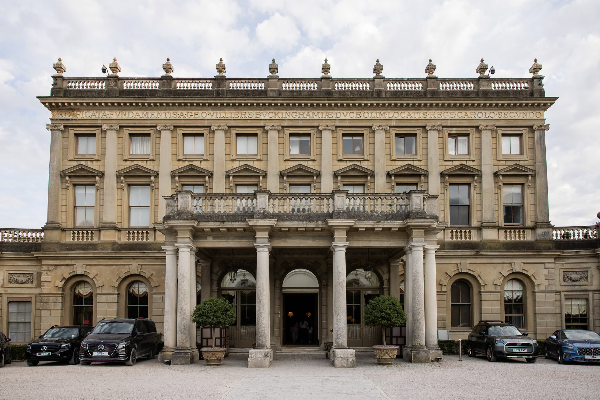 Grand historic mansion with stone columns, tall windows, and ornate architectural details. Four cars are parked in front, and two large potted plants flank the entrance. The sky is partly cloudy. David Bostock Photography