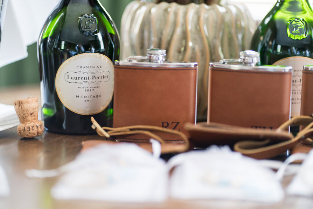 A bottle of Laurent-Perrier champagne, two brown leather hip flasks, a cork, and blurred objects on a table with a decorative background. David Bostock Photography