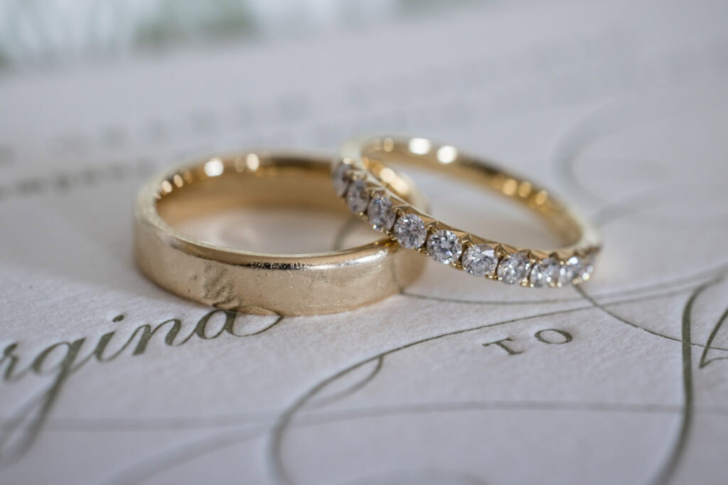 Two gold wedding rings rest on an elegant invitation; one ring is plain, and the other is adorned with diamonds. David Bostock Photography