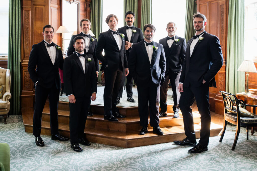 Eight men in black dinner jackets with bow ties pose together on wooden steps in a well-lit, elegant room with large windows and green curtains. David Bostock Photography