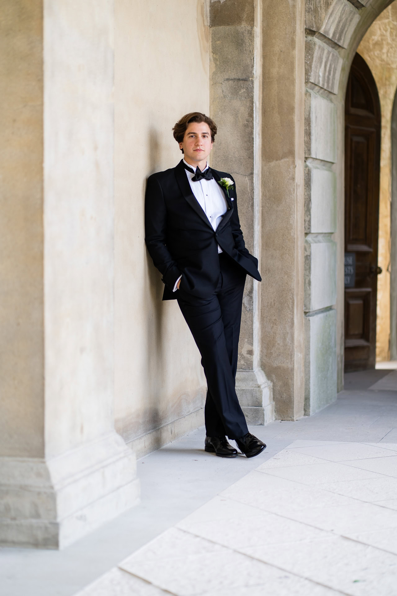 A young man in a black dinner jacket with a white shirt and bow tie leans against a stone wall under an archway, hands in pockets, looking at the camera. David Bostock Photography