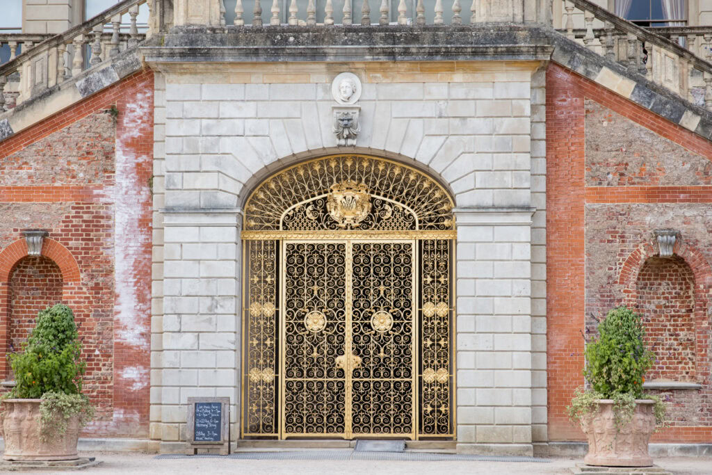 Ornate gold wrought iron gate set in a stone and red brick wall, flanked by arched alcoves with potted greenery, and a small blackboard sign near the entrance. David Bostock Photography