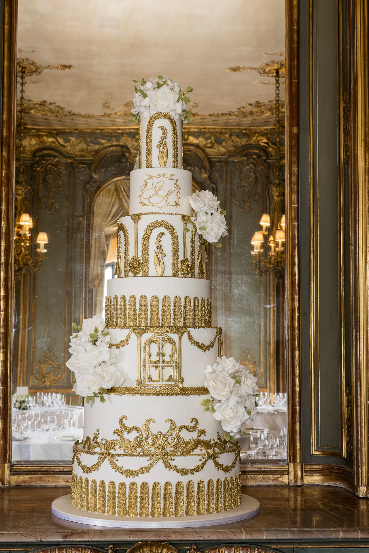 A tall, ornate white wedding cake with gold decorations and white flowers, displayed in front of a large, decorative mirror in an elegant, opulent room. David Bostock Photography