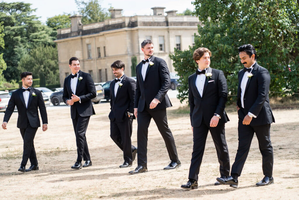 Six men in black dinner jackets with buttonholes walk together outdoors on a sunny day, with a large historic building and trees in the background. David Bostock Photography