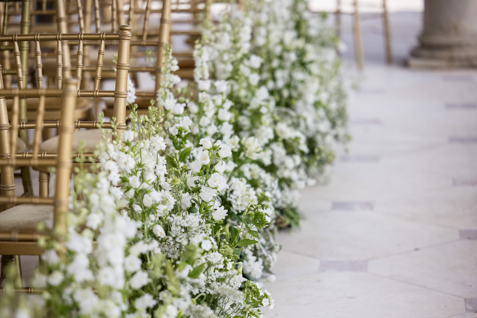 Rows of gold chairs line a stone aisle, which is decorated with lush arrangements of white flowers and greenery, creating an elegant and romantic setting, possibly for a wedding ceremony. David Bostock Photography