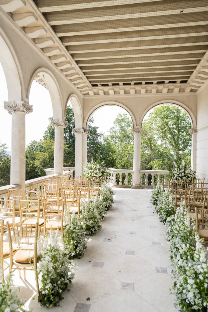 Elegant outdoor wedding ceremony setup with gold chairs and white floral arrangements lining the aisle, under a covered stone terrace with arches and tall columns overlooking green trees. David Bostock Photography