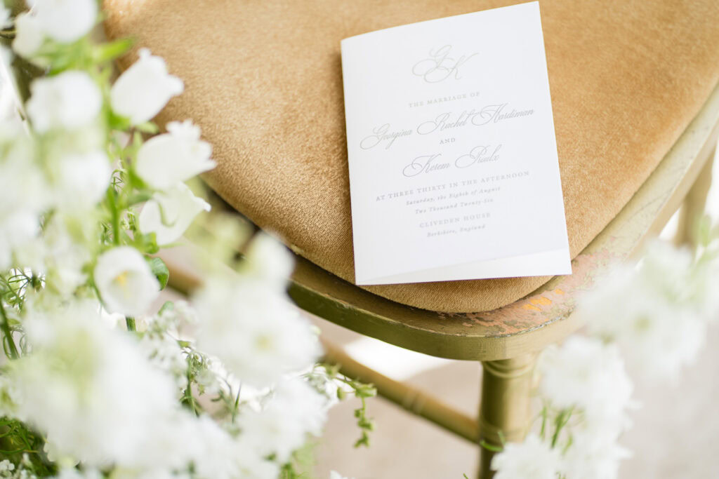A wedding invitation card rests on a gold cushioned chair surrounded by white flowers. The card features elegant script text with details about the marriage ceremony. David Bostock Photography