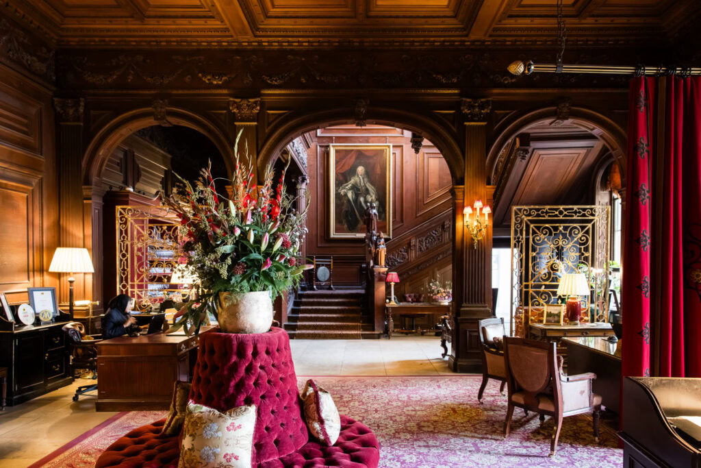 A luxurious, wood-panelled room with ornate arches, elegant furniture, a large floral arrangement, red carpet, and a grand staircase with a portrait hanging above it. Warm lighting enhances the opulent atmosphere. David Bostock Photography