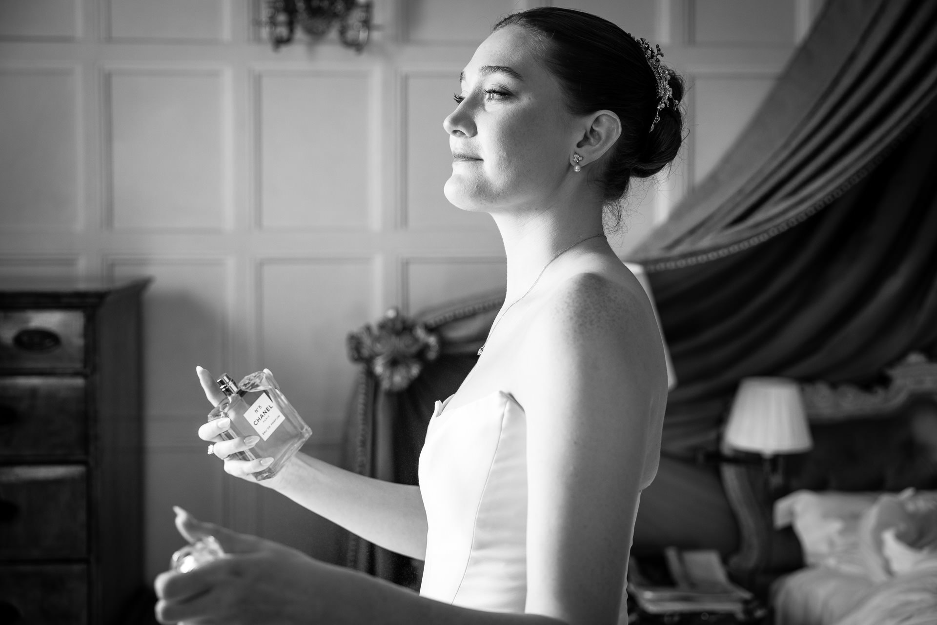 A bride in a white dress sprays perfume on herself whilst standing in a decorated room, smiling softly. David Bostock Photography
