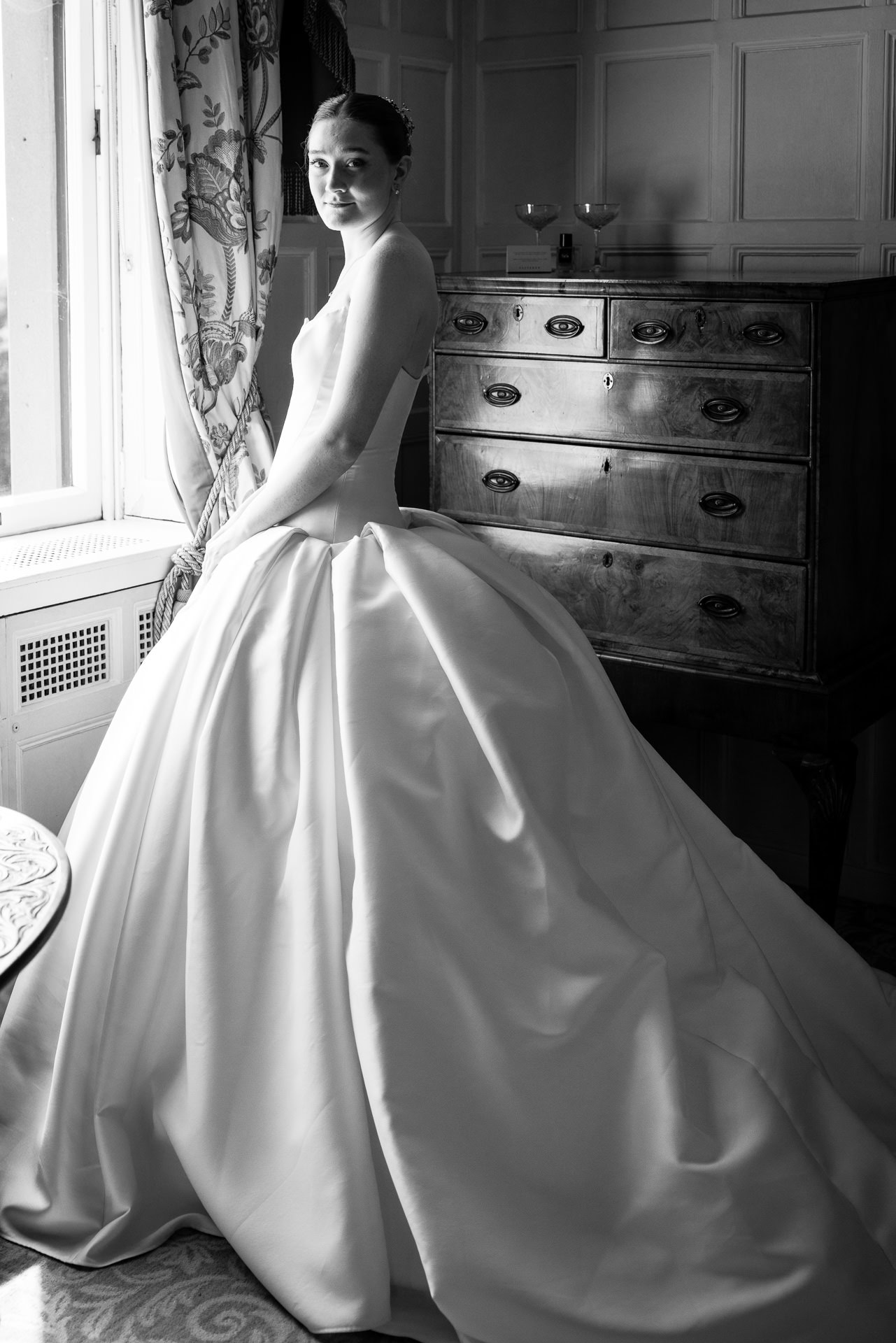 A woman in a strapless wedding dress sits beside a window, looking towards the camera. Light from the window illuminates her and the room, which features a wooden chest of drawers and patterned curtains. David Bostock Photography