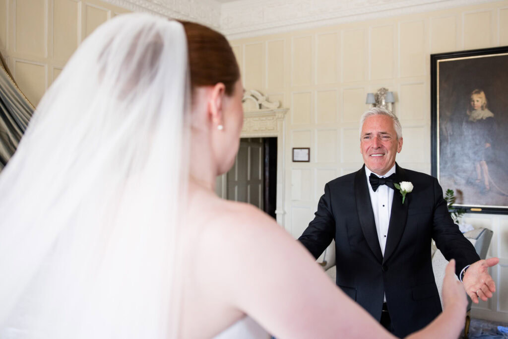 A bride in a wedding dress and veil faces an older man in a tuxedo with a white rose buttonhole, who stands with open arms and an emotional expression in an elegant room. David Bostock Photography