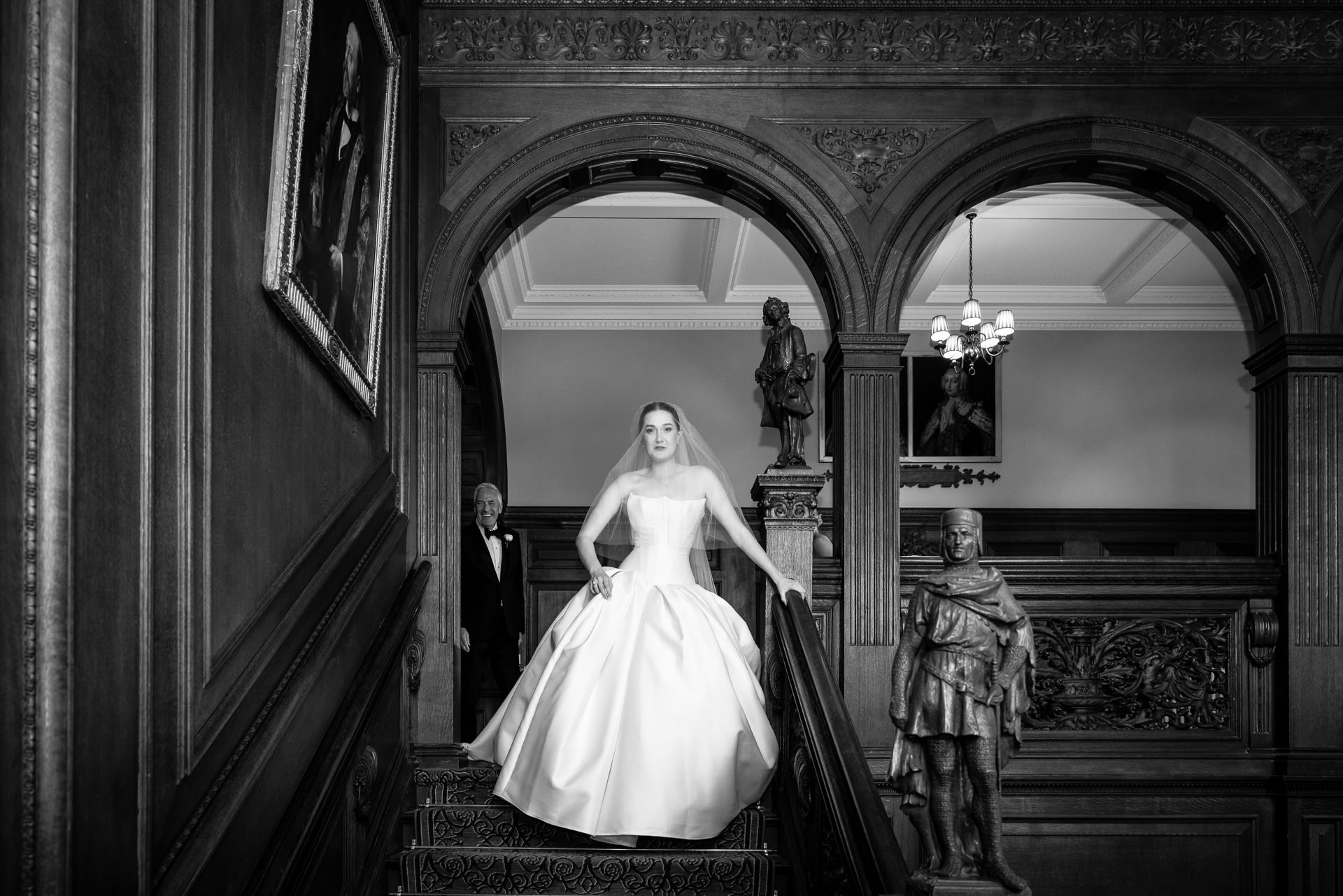 A bride in a white dress walks down an ornate staircase, with an older man behind her. The grand interior features statues, arches, wood panelling, and framed portraits. David Bostock Photography