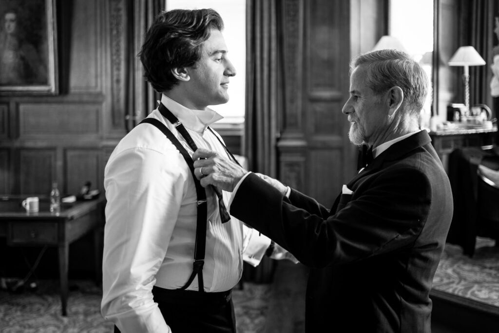 A man adjusts another man's bow tie in an elegant, wood-panelled room. Both wear formal attire and smile warmly, suggesting a special occasion or celebration. David Bostock Photography