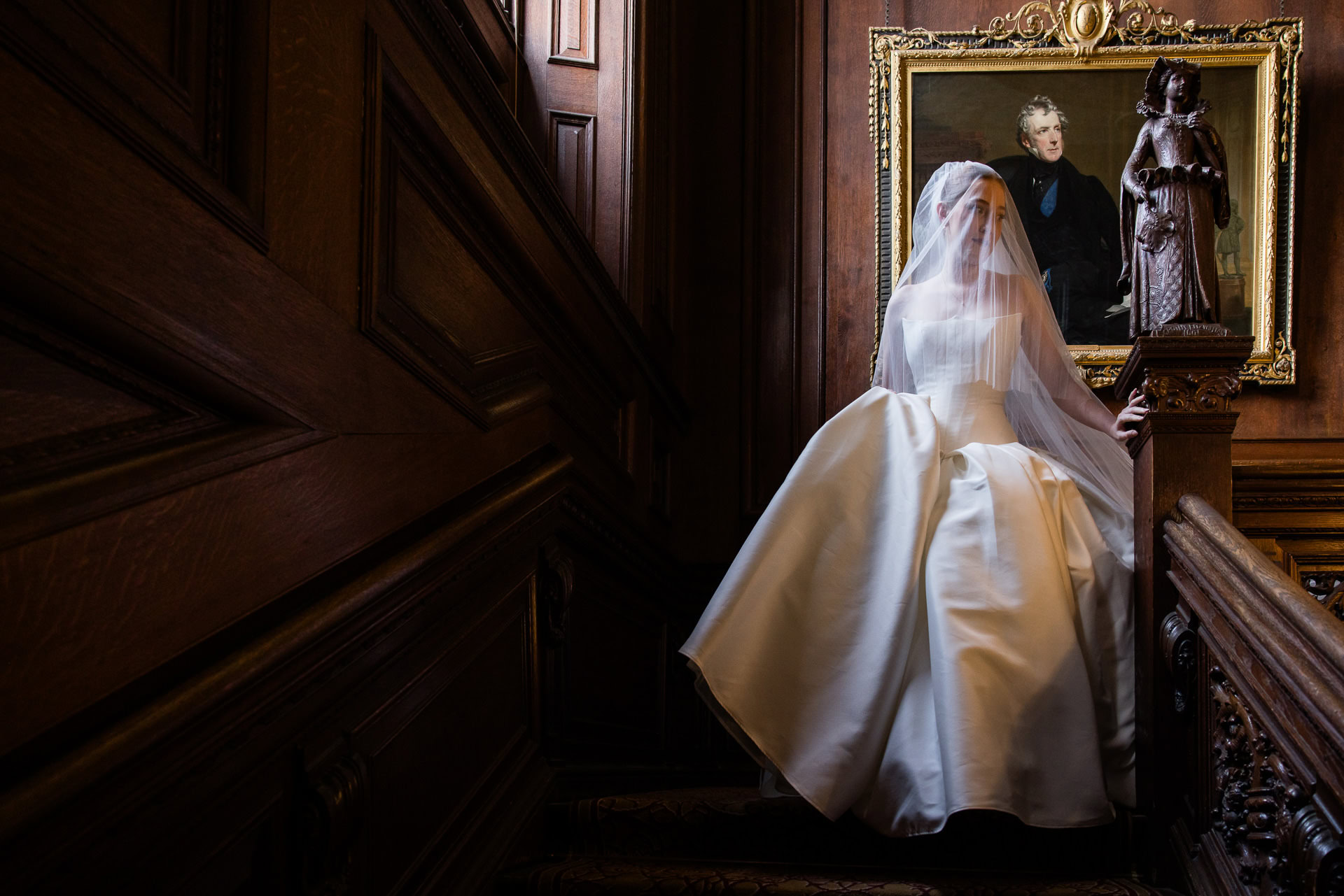 A bride in a white dress and veil descends a wooden staircase, with a framed portrait hanging on the panelled wall behind her. David Bostock Photography