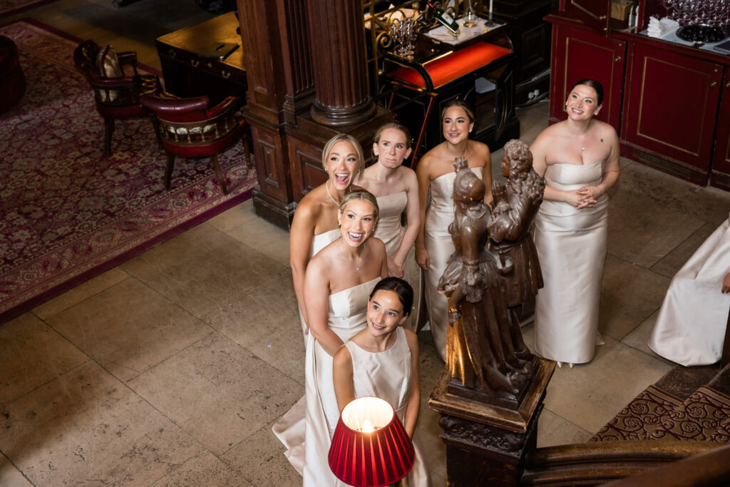 Six women in matching off-white dresses stand on a staircase, smiling and looking up. The setting appears elegant, with wood panelling, red carpet, and ornate decor, including statues and a red lamp. David Bostock Photography