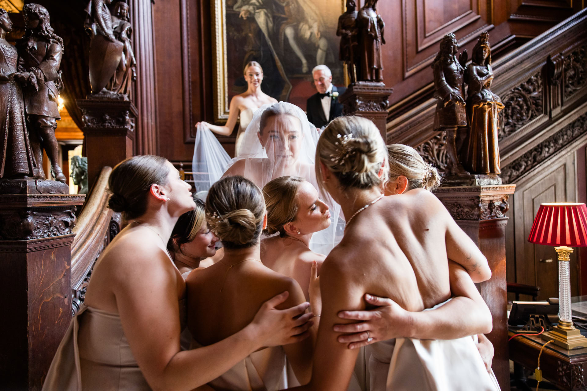 A bride in a veil stands on a grand staircase, surrounded by bridesmaids in light dresses, embracing her. Two people stand above, smiling, in an ornate, wood-panelled room with statues and a red lamp. David Bostock Photography