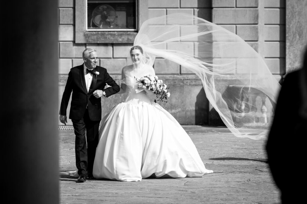 A bride in a flowing gown and veil walks arm-in-arm with an older man in a tuxedo outside a stone building, both smiling. David Bostock Photography