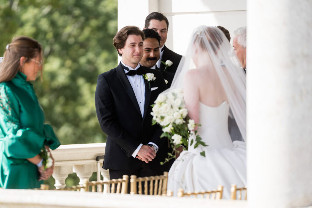 A groom in a black tuxedo stands facing a bride in a white gown and veil, holding a bouquet. Others stand nearby, including a woman in green, during an outdoor wedding ceremony. David Bostock Photography