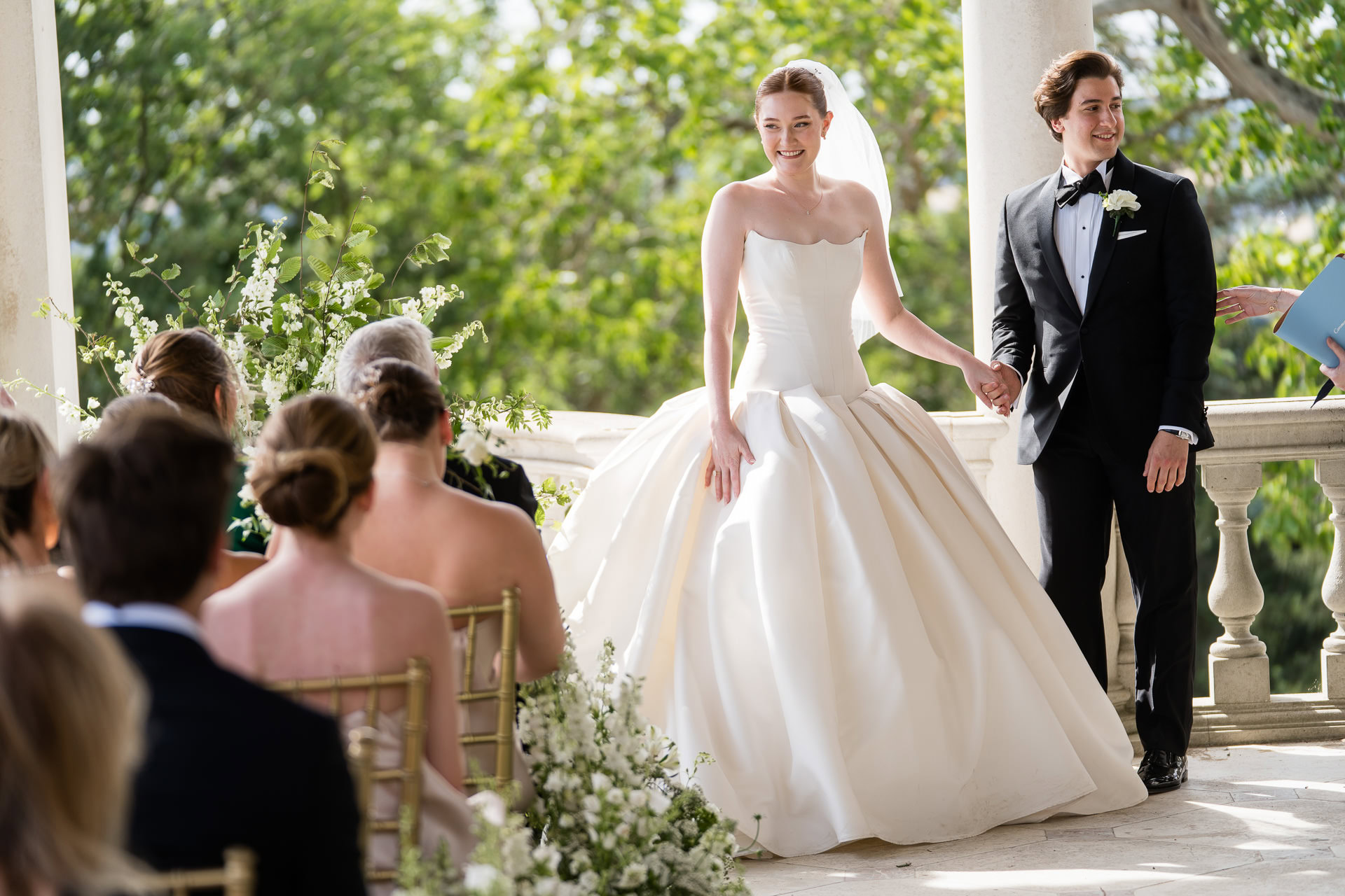 A bride and groom stand smiling at an outdoor wedding ceremony, holding hands in front of seated guests. The bride wears a white gown and veil; the groom is in a black dinner suit. Greenery and flowers decorate the scene. David Bostock Photography
