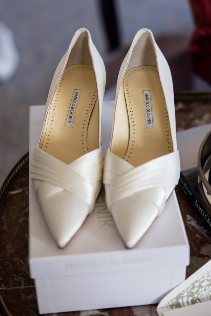 A pair of white Manolo Blahnik high-heeled shoes with pointed toes sits atop a white shoebox on a marble surface. David Bostock Photography