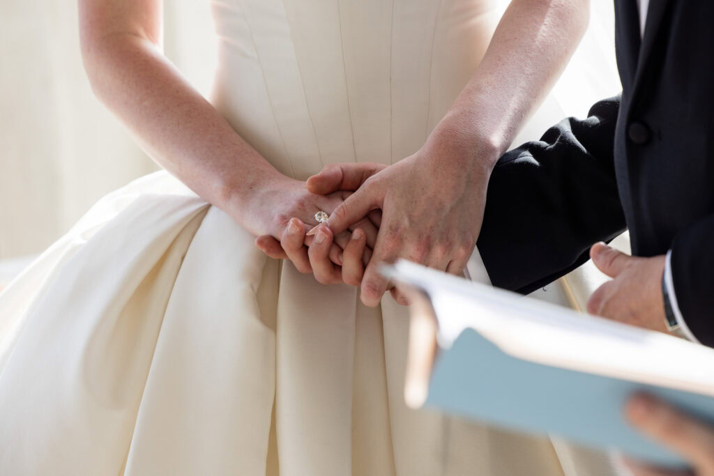 Close-up of a bride and groom holding hands during a wedding ceremony, with the bride in a white dress and the groom holding a book, possibly a vow book or script. David Bostock Photography
