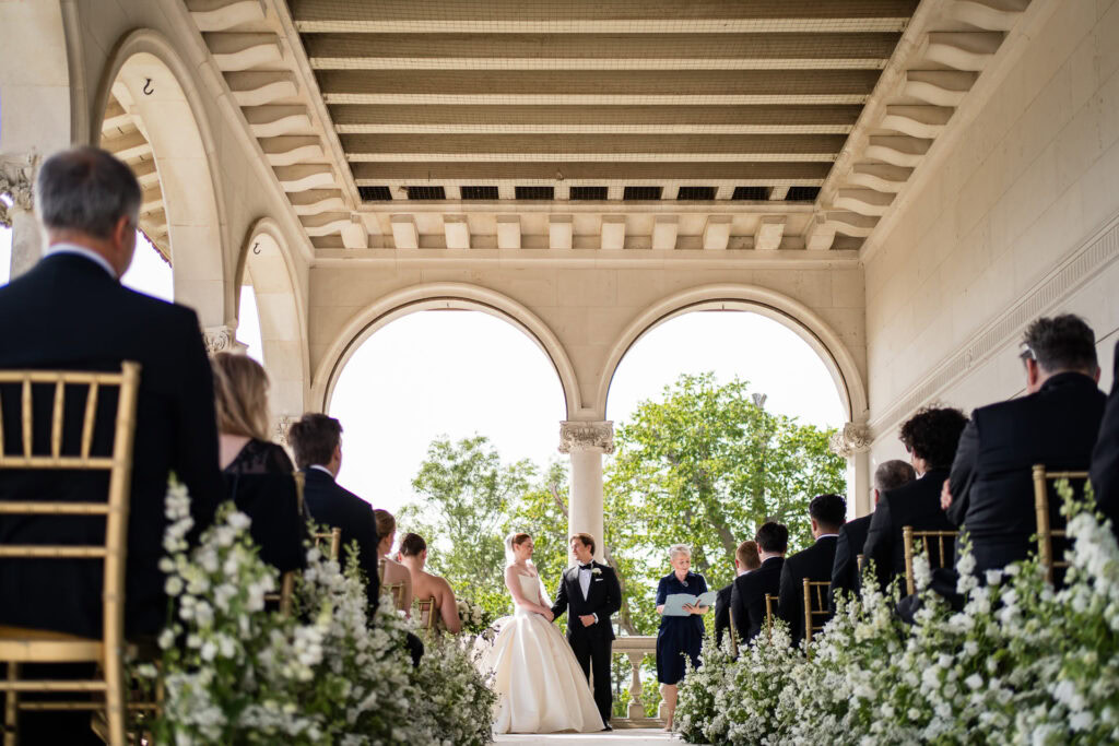 A bride and groom stand at the altar during an outdoor wedding ceremony, surrounded by guests seated on gold chairs under a vaulted pavilion decorated with white flowers. David Bostock Photography