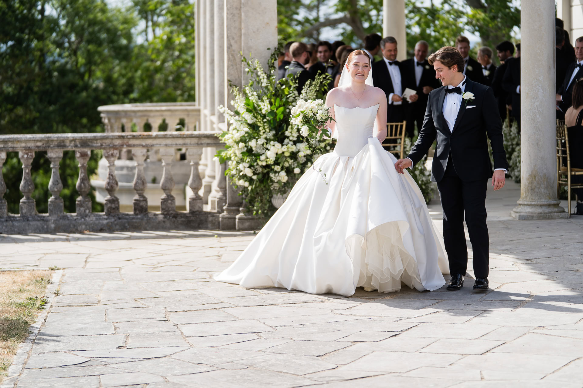 A bride in a white gown and a groom in a black tuxedo walk outside at a sunny wedding ceremony, surrounded by greenery, columns, and seated guests in formal attire. David Bostock Photography