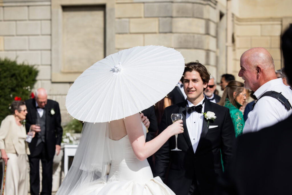 A bride holding a white parasol talks to a man in a tuxedo at an outdoor wedding, with guests mingling in the background. David Bostock Photography