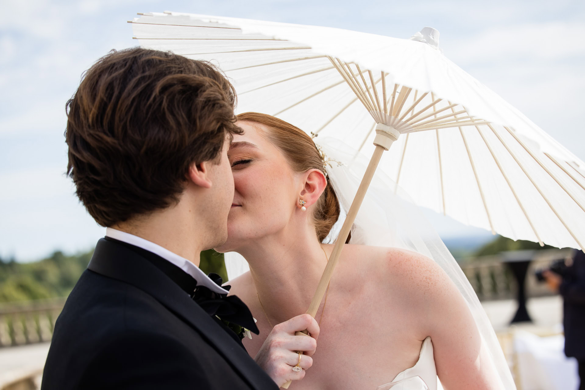 A bride and groom share an intimate moment under a white parasol, with the bride leaning in to kiss the groom outdoors on a sunny day. David Bostock Photography