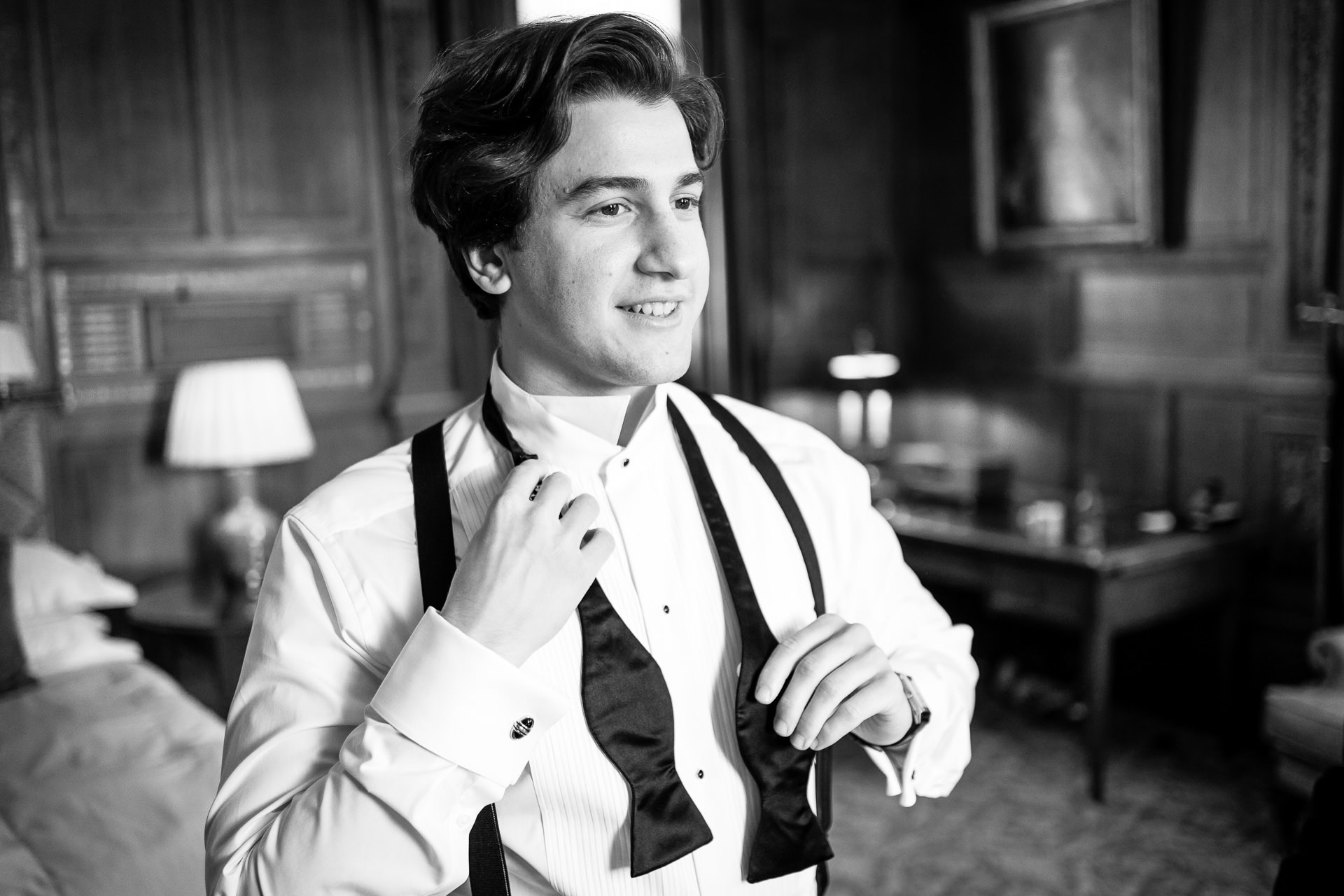 A young man in formal attire stands in a room, smiling whilst adjusting his bow tie. The room features wooden panelling, a lamp, and framed art. The image is in black and white. David Bostock Photography