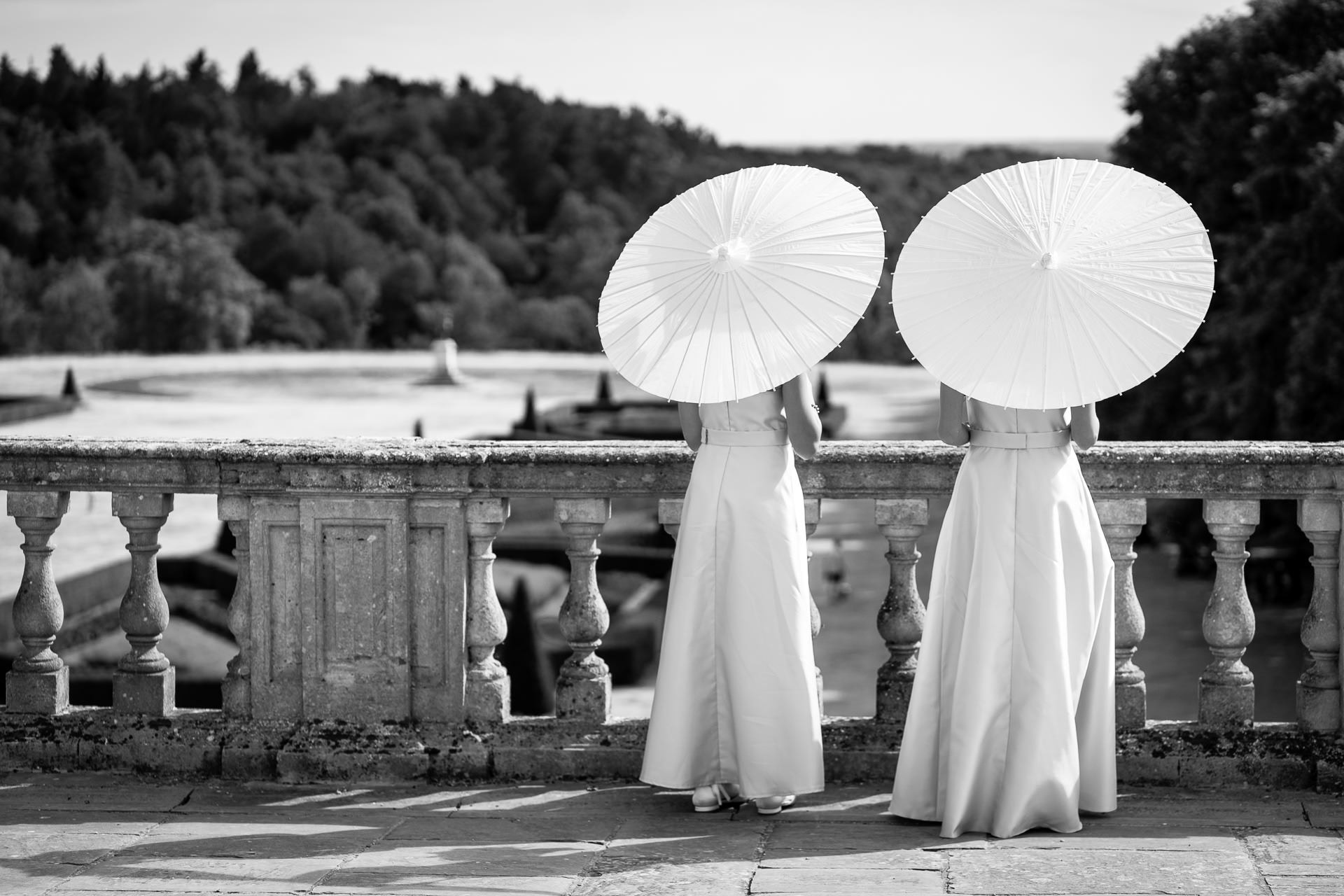 Two women in long dresses stand by a stone balustrade, each holding a white parasol. They face away, overlooking a scenic, tree-filled landscape. The photo is in black and white. David Bostock Photography