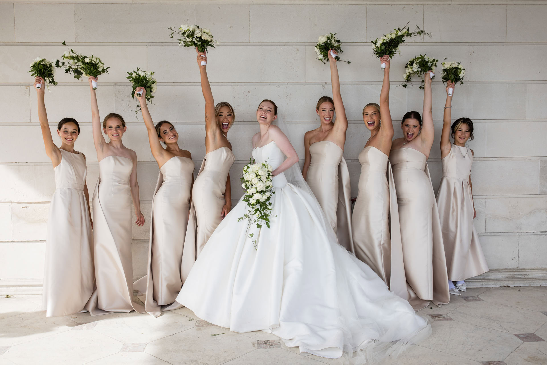 A bride in a white dress stands with seven bridesmaids in light dresses, all smiling. The bridesmaids hold bouquets aloft, celebrating together against a light stone wall background. David Bostock Photography