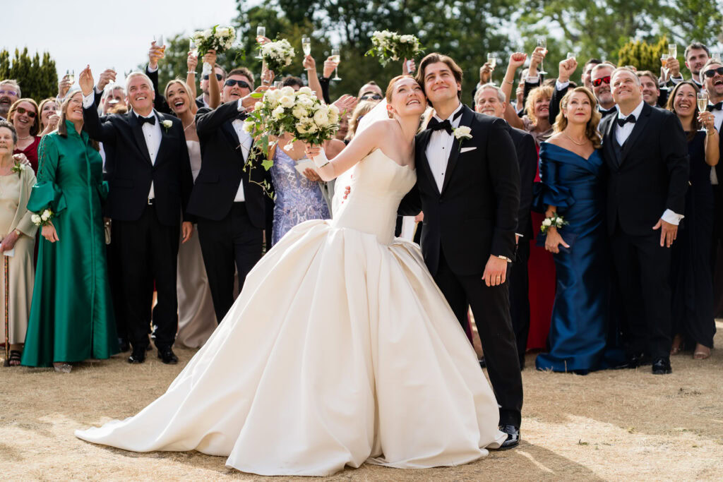 A bride and groom smile and pose in front of a large group of well-dressed guests, who are raising glasses in celebration outdoors on a sunny day. David Bostock Photography