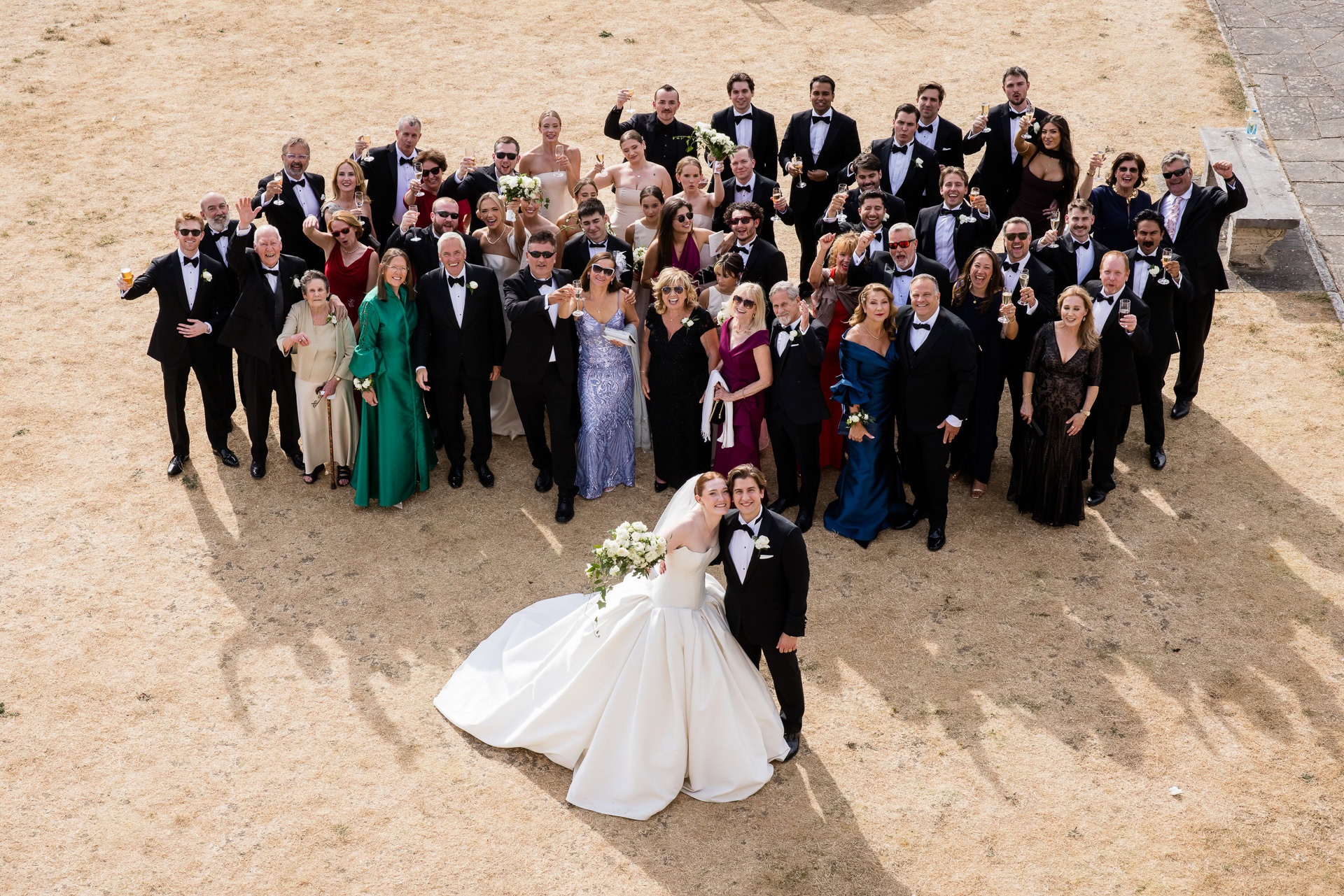 A bride and groom kiss in front of a large group of guests dressed formally, posing together outdoors on a patchy, dry grassy area. David Bostock Photography