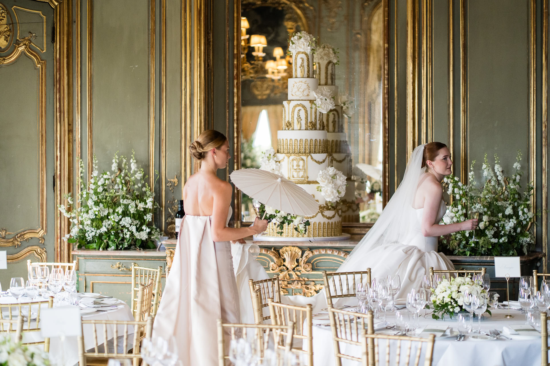 Two women in elegant dresses stand in a lavishly decorated room with ornate walls, gold trim, white floral arrangements, and a tall tiered wedding cake. One woman holds a parasol. Tables are laid for a formal event. David Bostock Photography