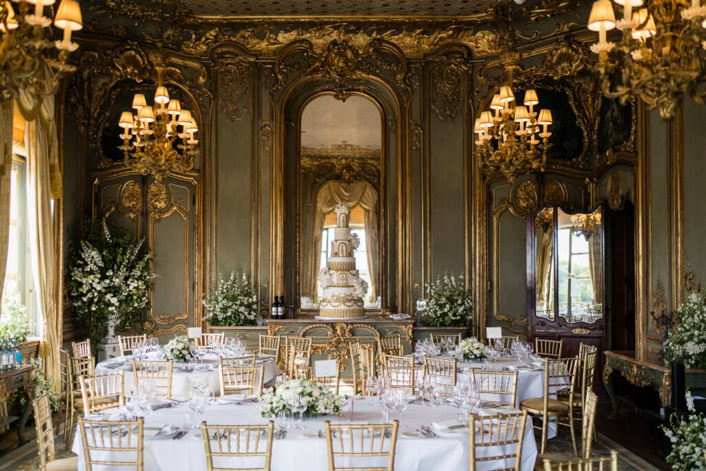 Elegant ballroom with ornate gold decor, round tables set for an event, gold chairs, floral centrepieces, chandeliers, and a large tiered wedding cake on display in front of a grand mirror. David Bostock Photography
