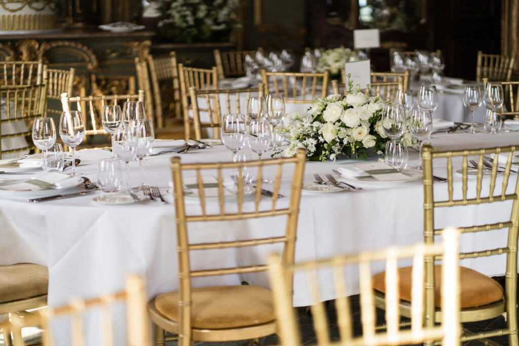 Elegant banquet table set with white tablecloth, glassware, cutlery, and a floral centrepiece, surrounded by gold chairs in a decorated room. David Bostock Photography