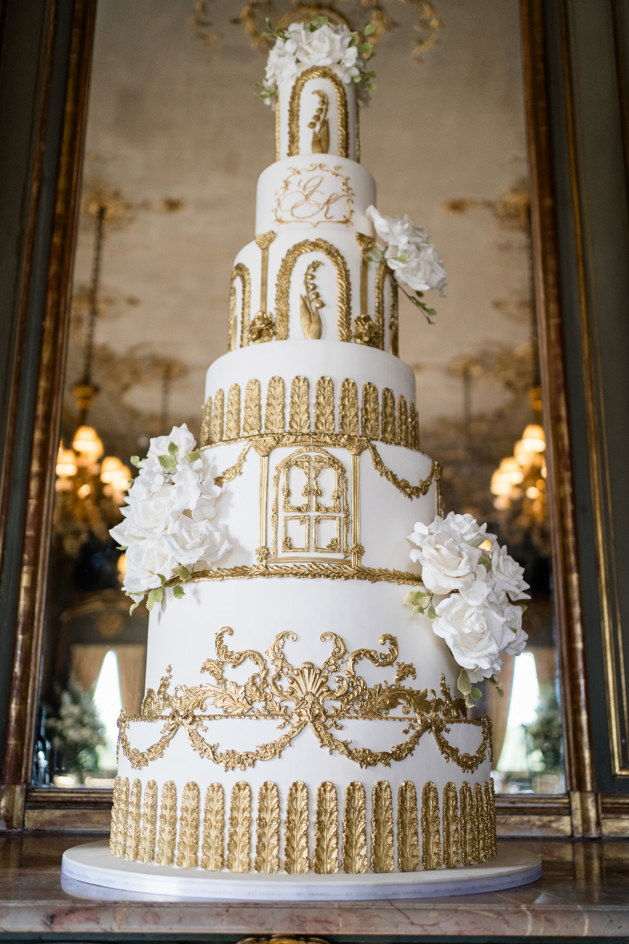 Tall, ornate white and gold tiered cake decorated with intricate gold designs and white flowers, reflected in a mirror behind it. David Bostock Photography